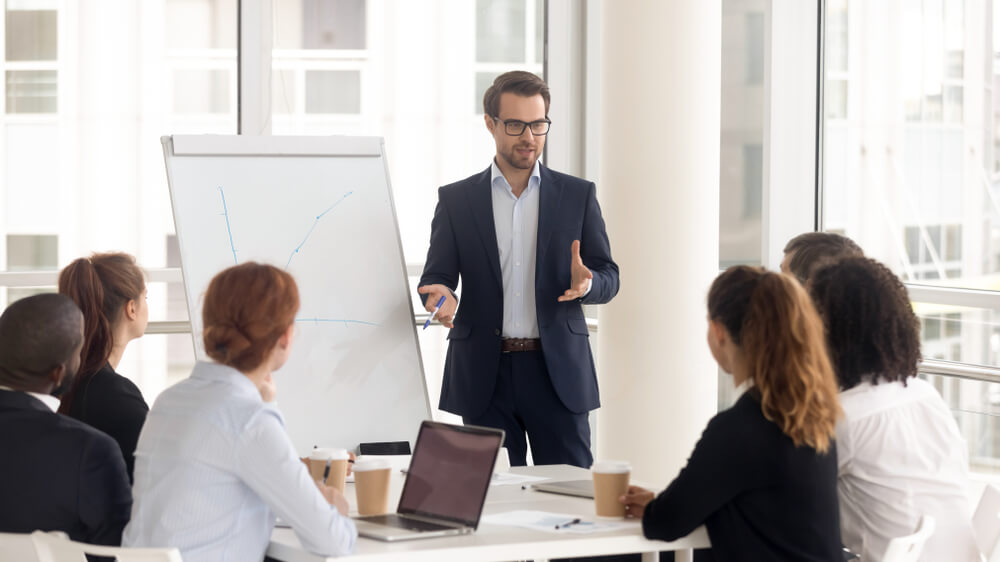 In a modern office setting, a man in a suit exudes leadership as he presents to a group seated around the table. Gesturing toward a blank flip chart, the team listens attentively, with laptops and coffee cups scattered about.